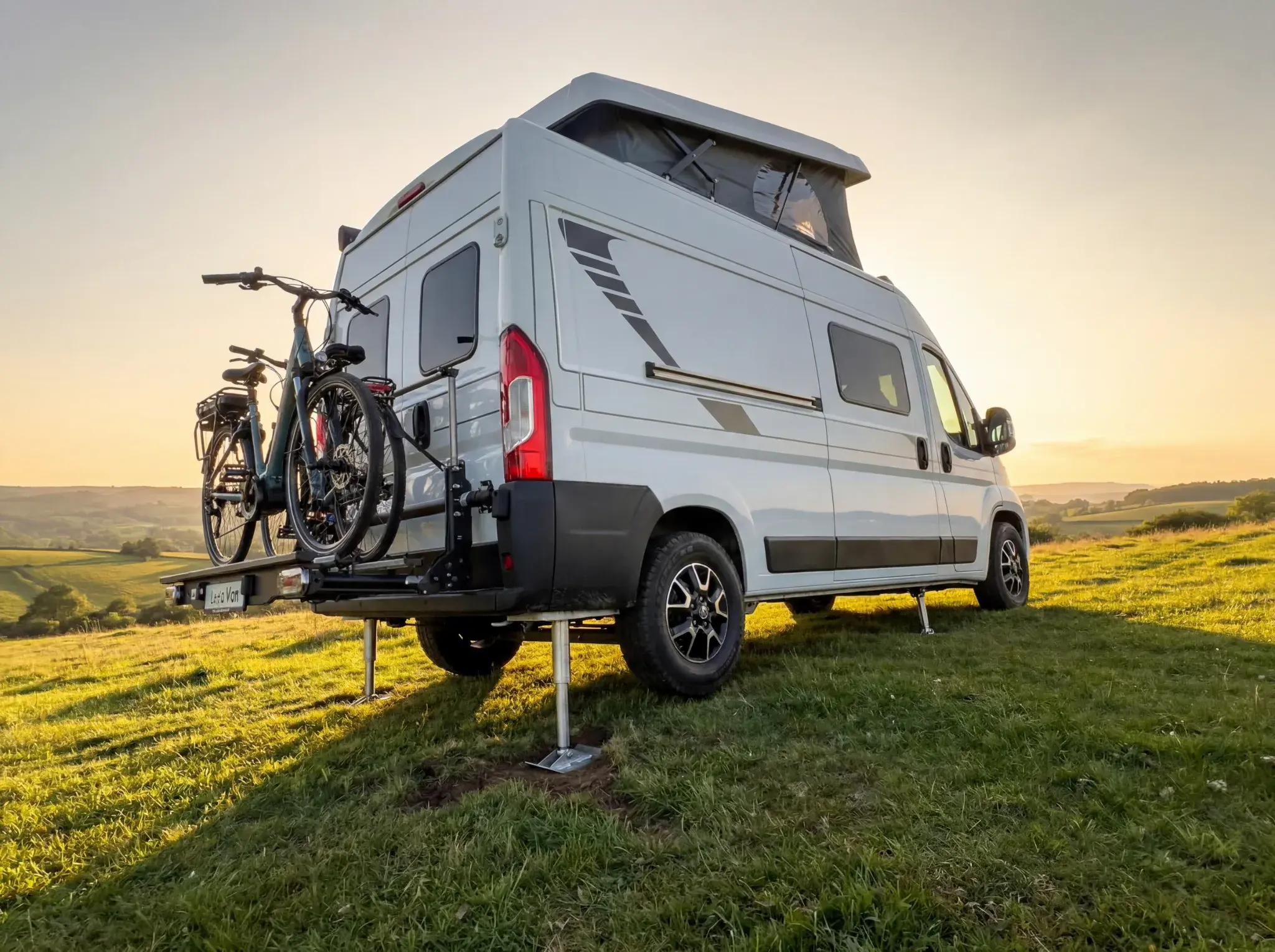 White compact campervan with raised pop-top roof parked on grass hillside, bicycles mounted on rear carrier.