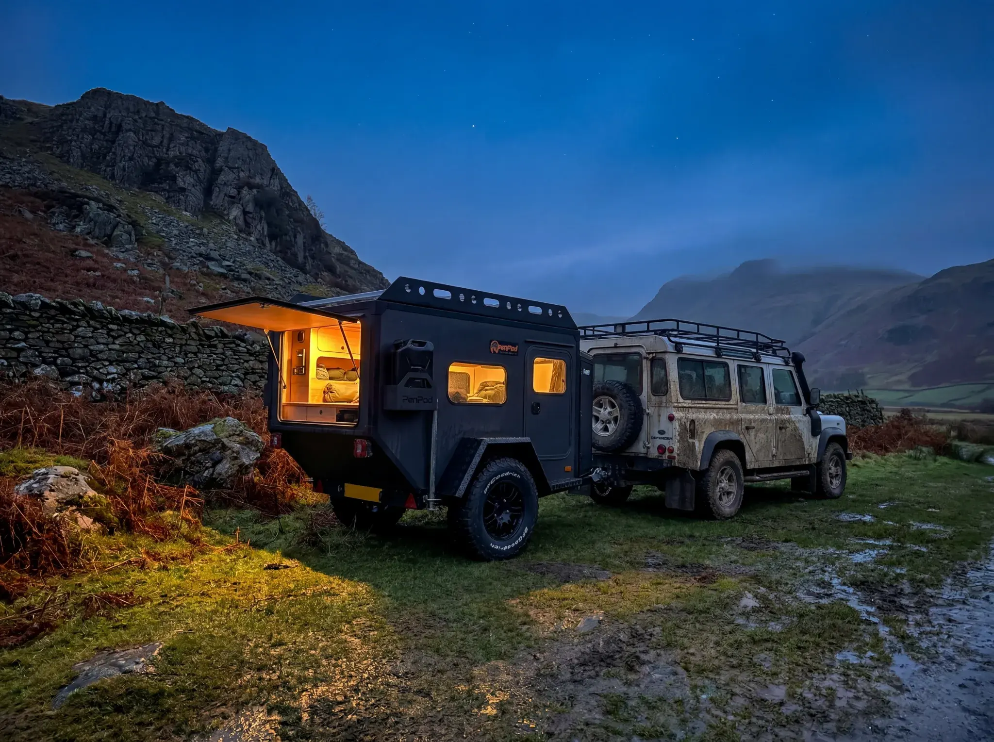 Black teardrop campervan with open orange-lit door towed behind white Land Rover Defender in mountain landscape at dusk.