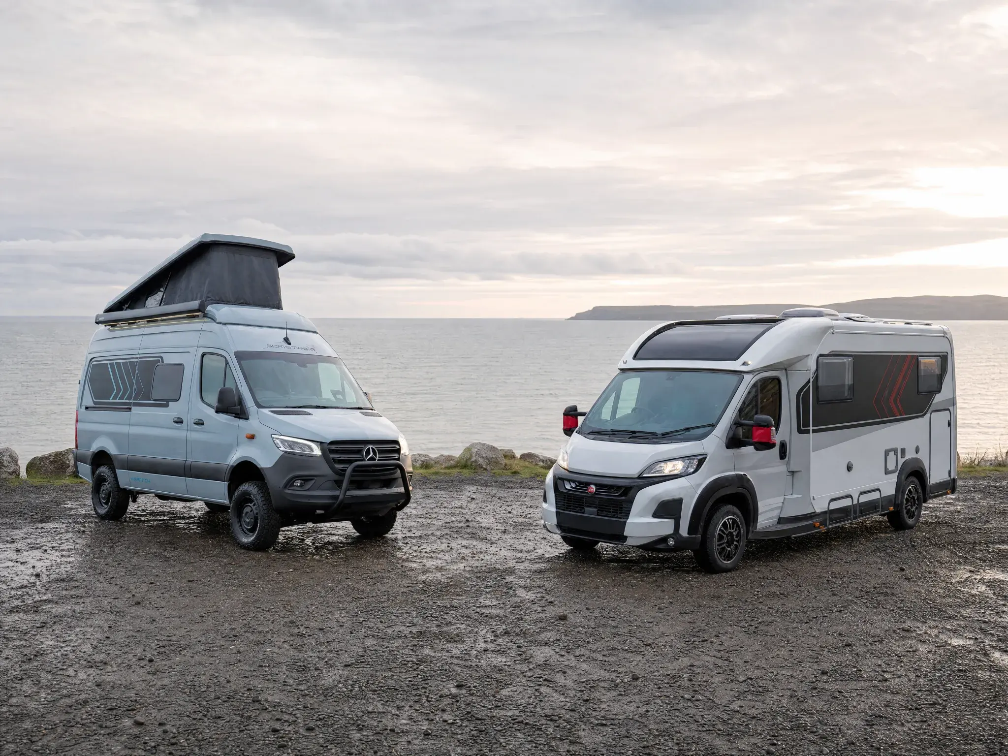 Two white campervans parked on gravel beach with ocean and coastal cliffs in background.