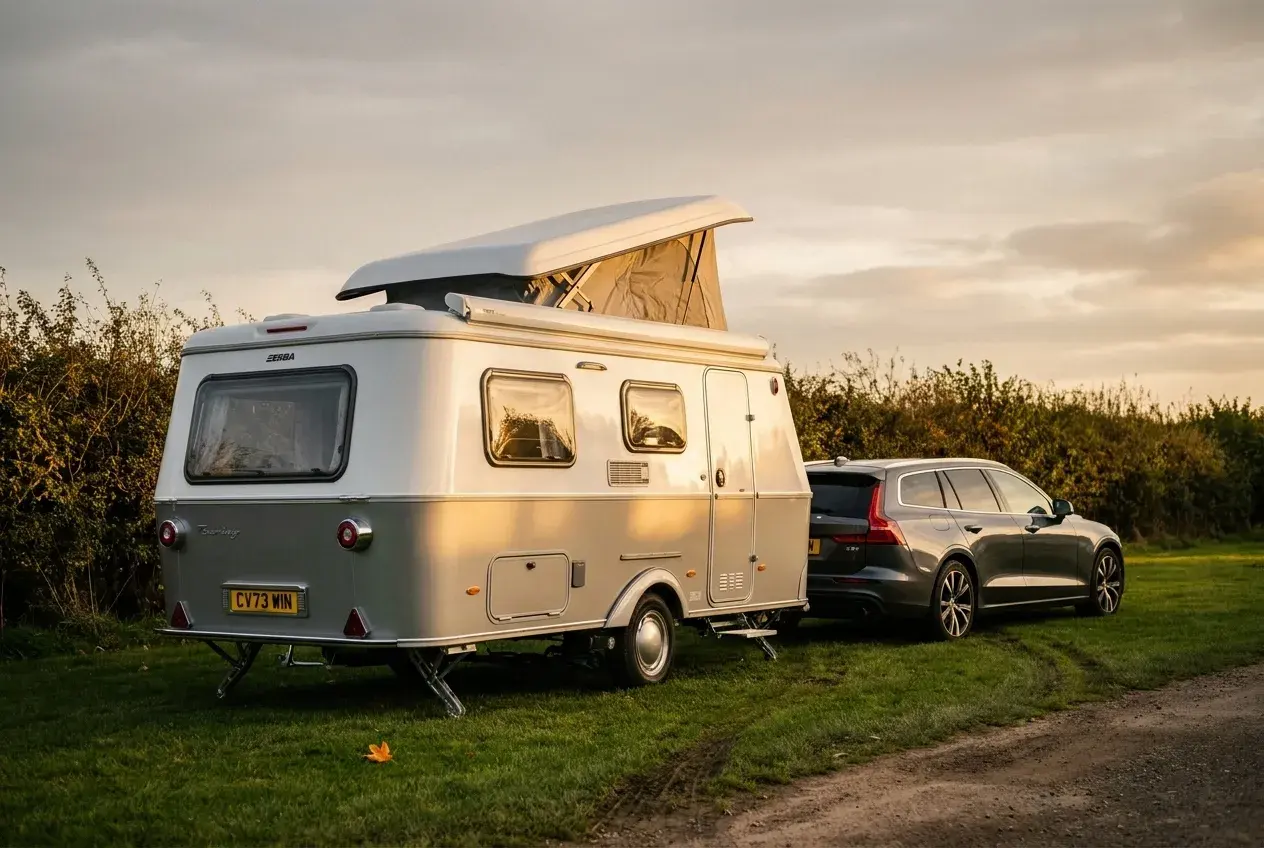 An Eriba pop-top touring caravan with its roof raised, parked beside a car on a grassy UK campsite pitch in warm evening light.