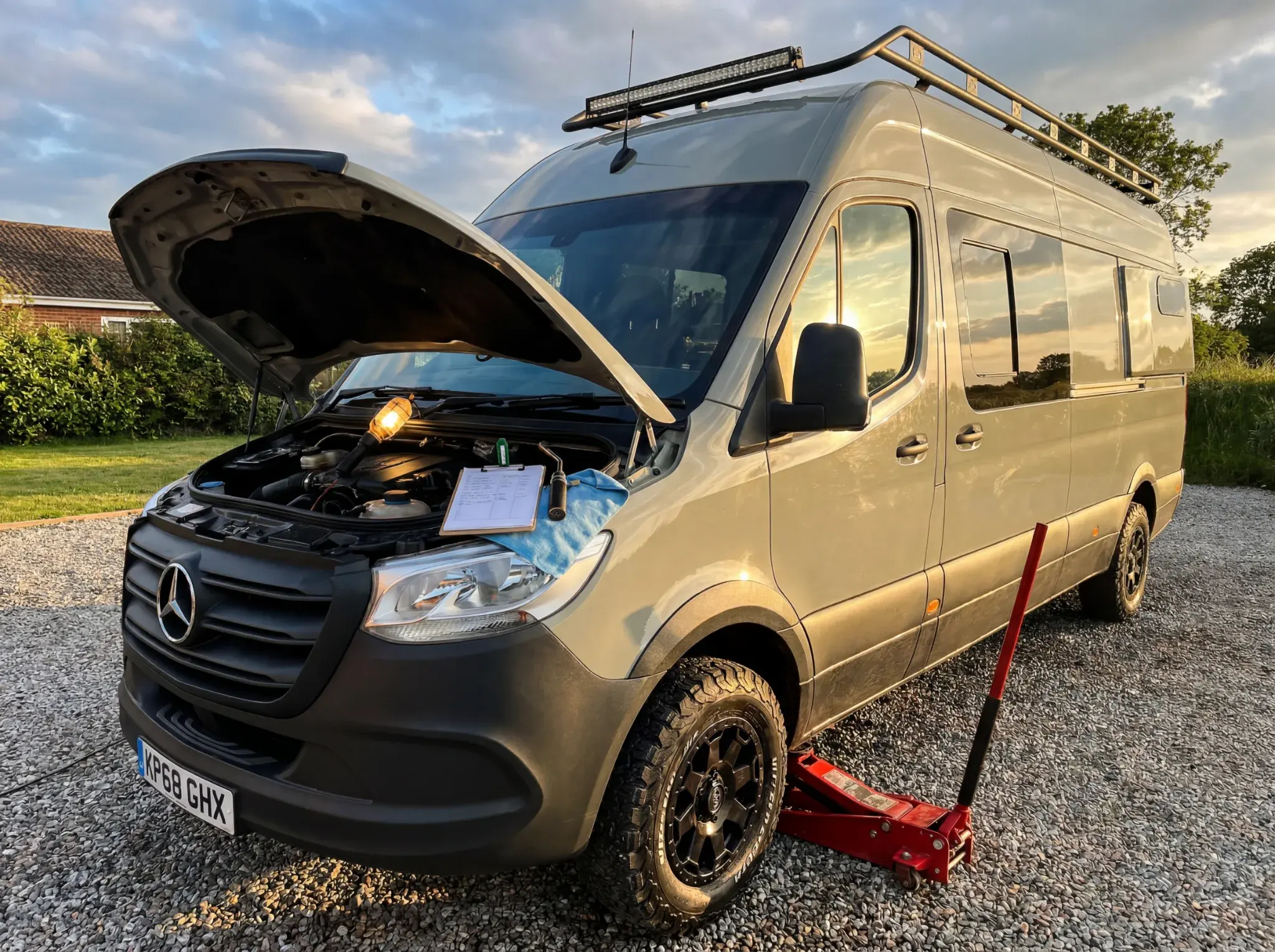 Silver Mercedes campervan with bonnet open, roof rack and ladder visible, parked on gravel.