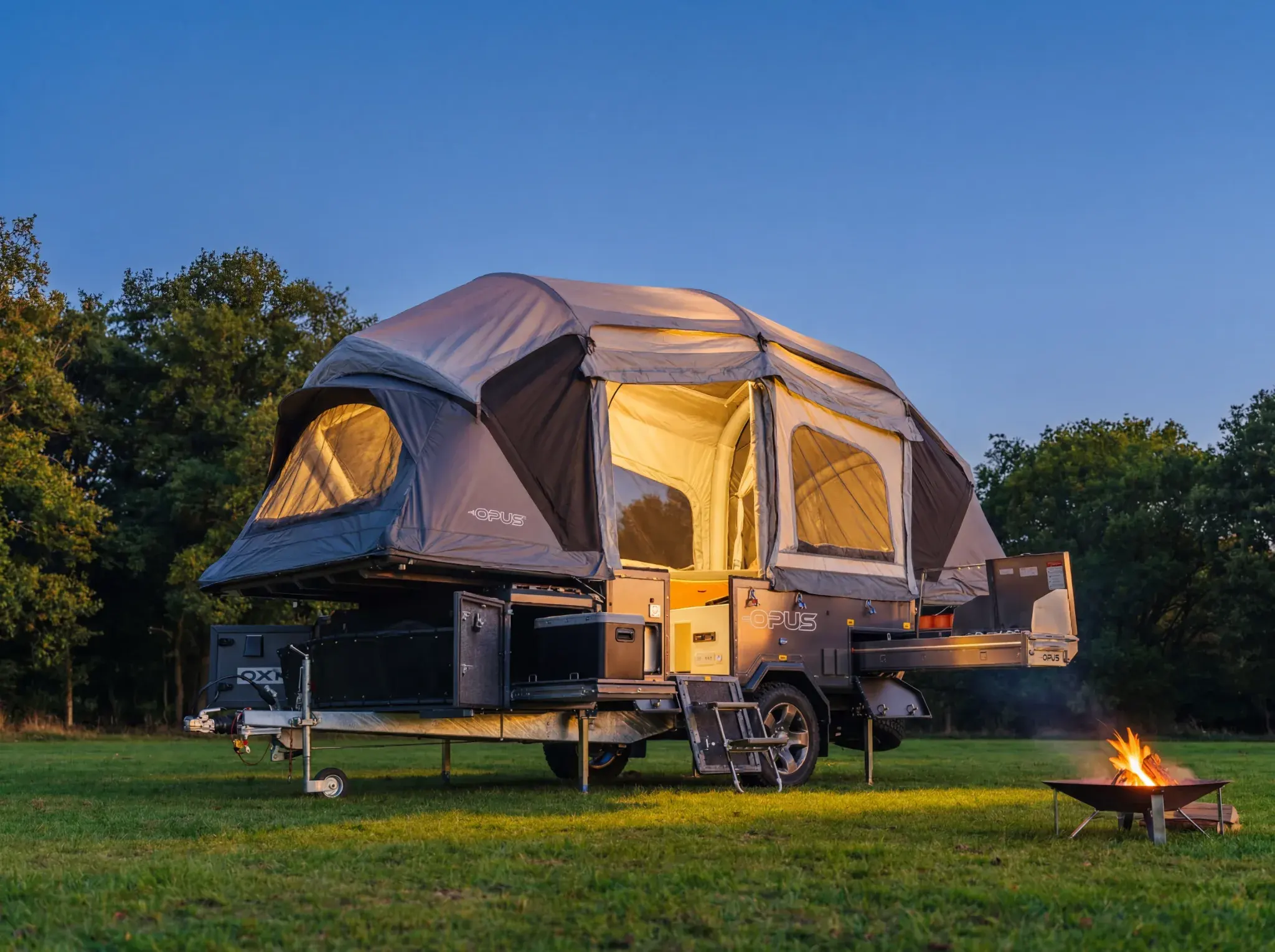 OPUS folding camper trailer with expanded tent body illuminated at dusk, campfire in foreground, parked on grass with trees behind.