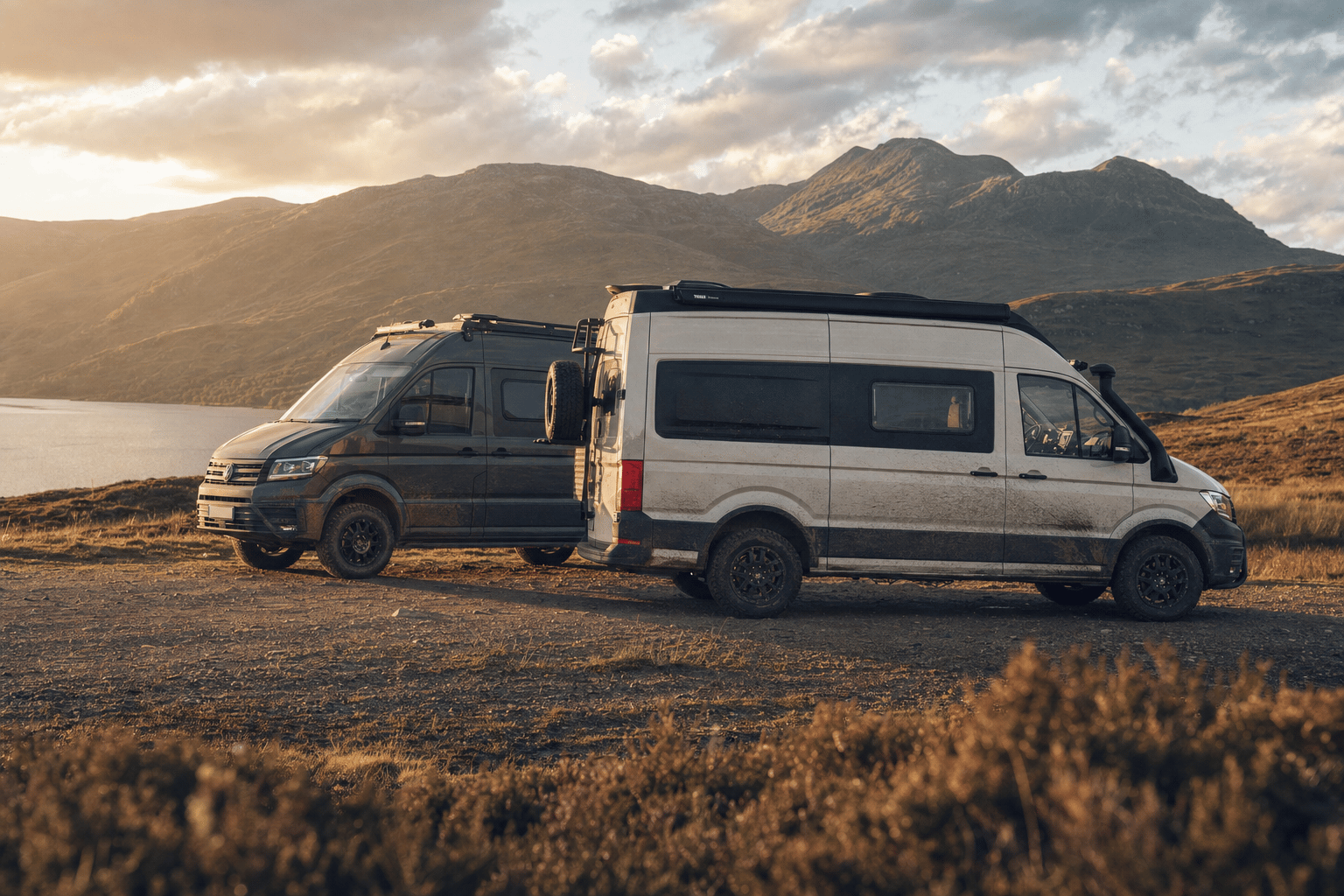 Two rugged 4x4 campervans, one smaller and one larger, parked on a remote Highland track at golden hour beside heather and hills.