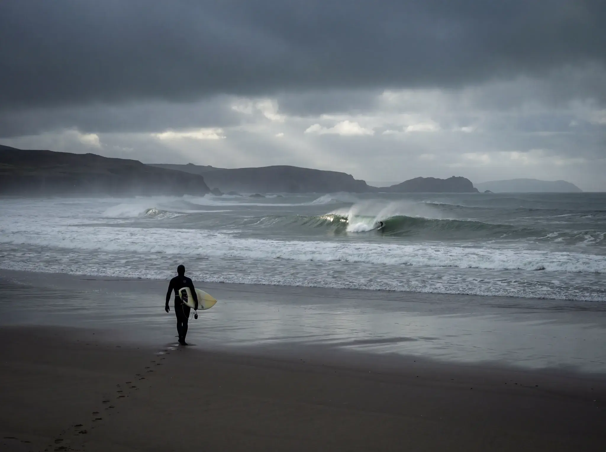 Surfer in black wetsuit carrying yellow board walking into ocean with rolling waves and rocky cliffs.