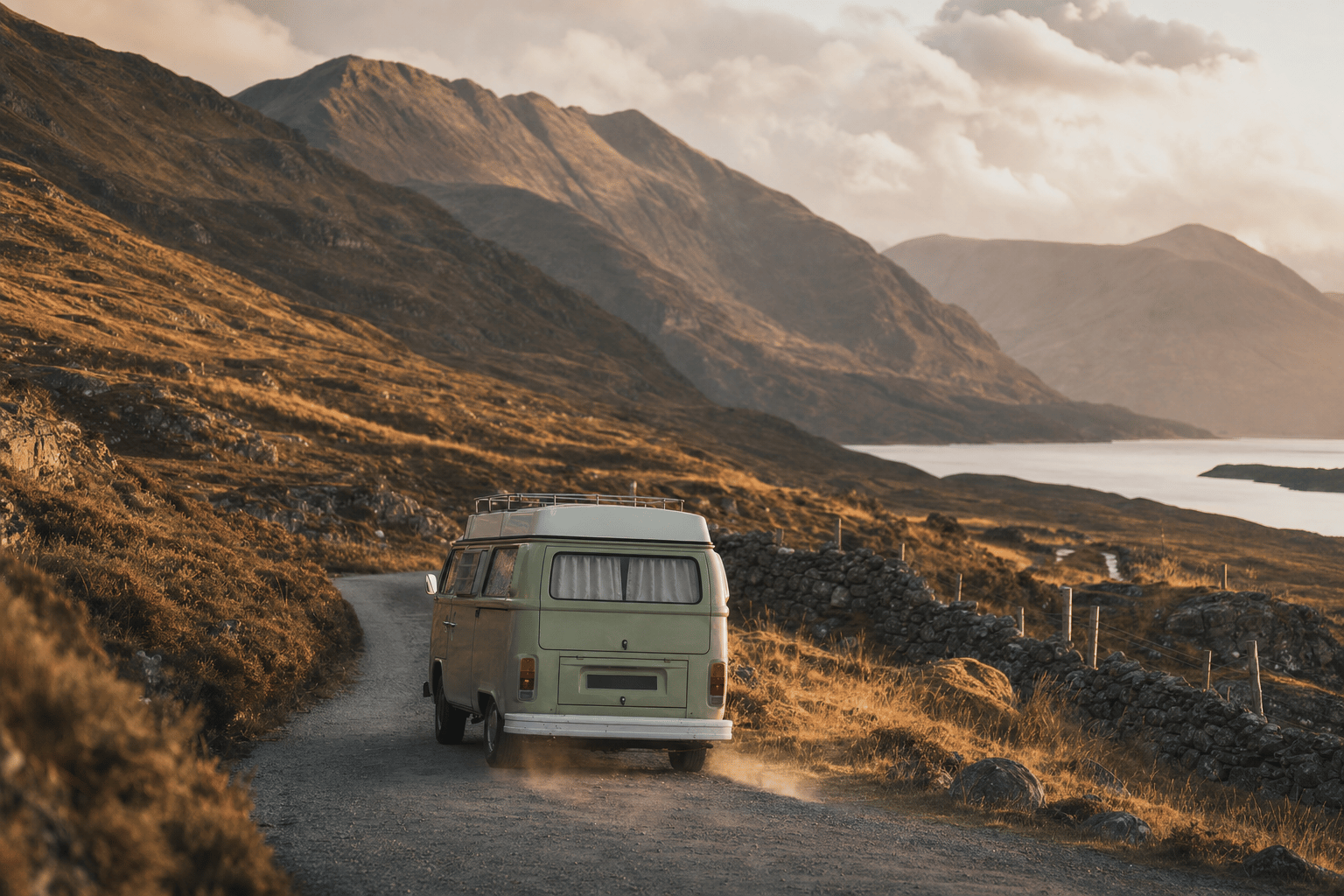 A campervan driving along a narrow winding road through Highland mountains and a loch in warm evening light.