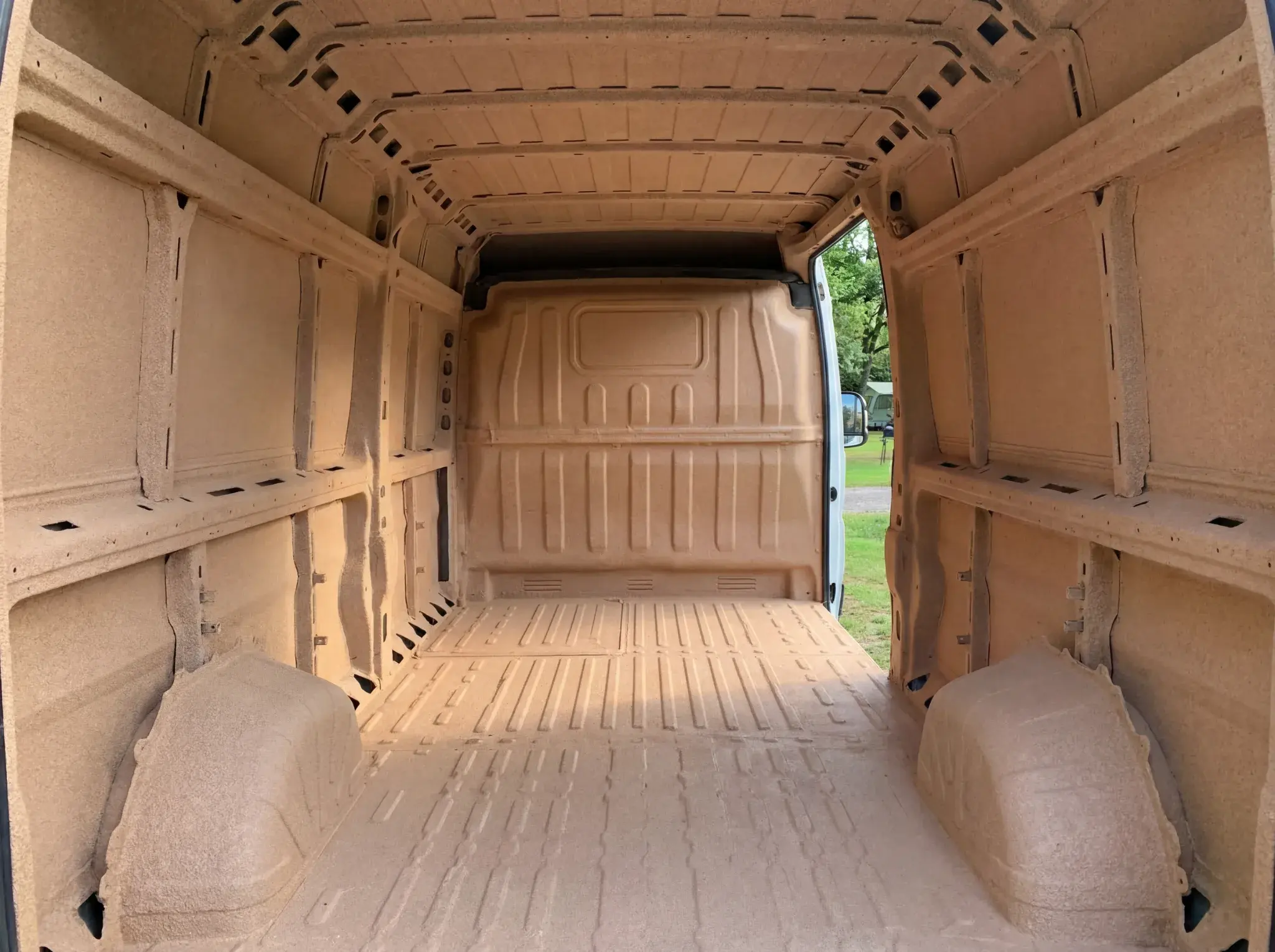 Empty van interior with beige walls, ribbed ceiling, shelving units on both sides, and open rear doors.