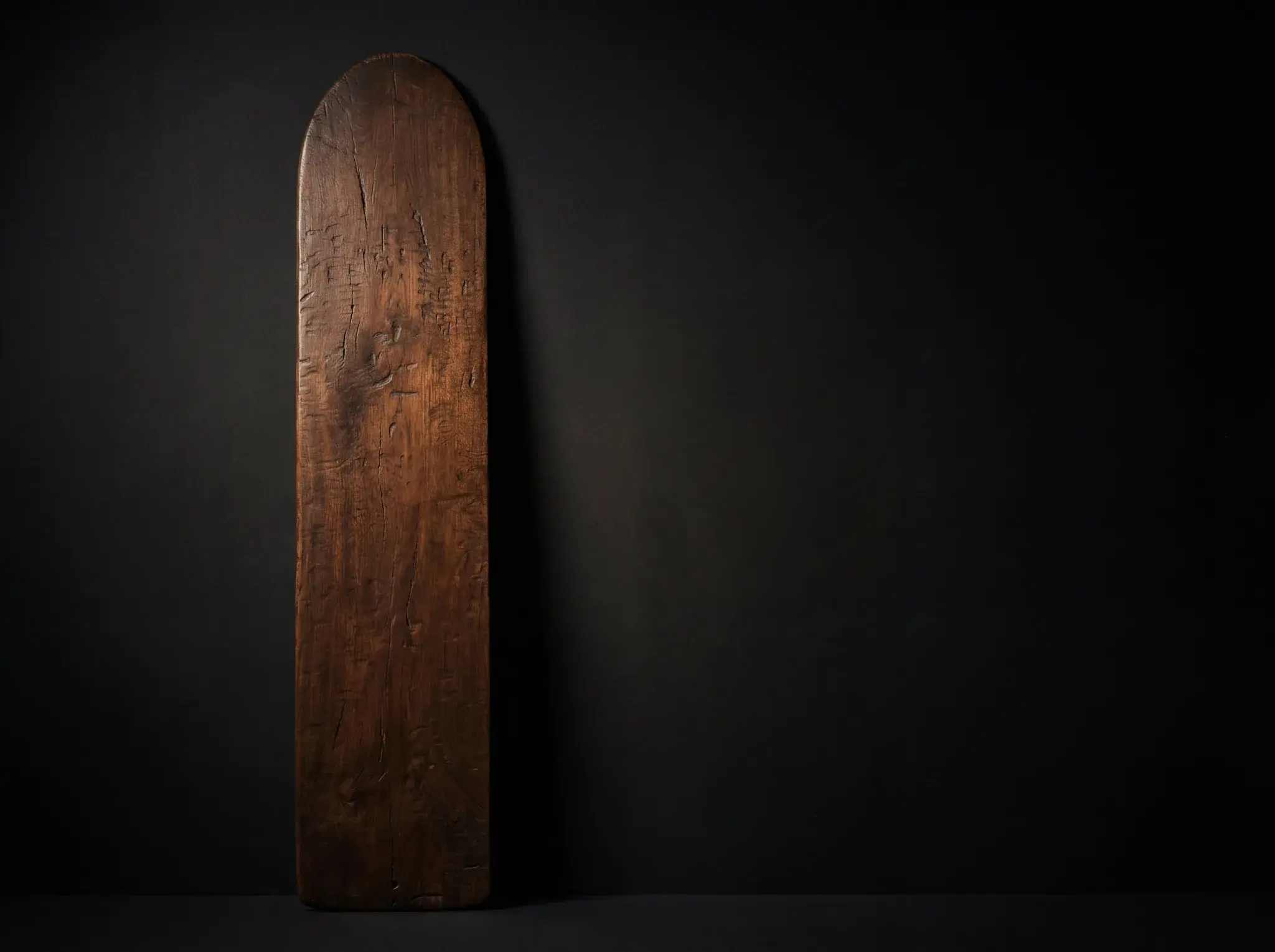 Antique wooden surfboard with carved details leaning against dark background.