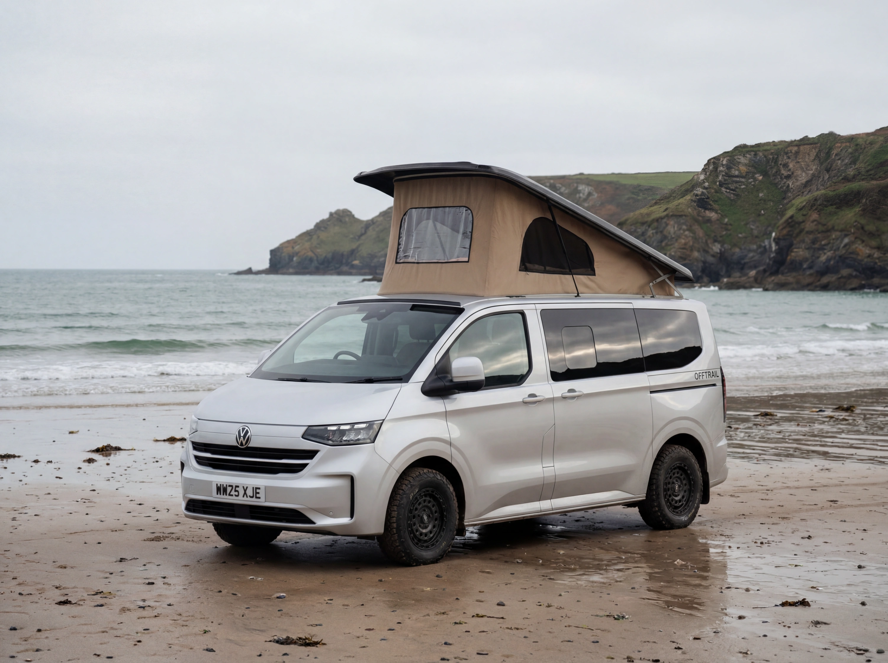 White Volkswagen van with tan pop-up roof parked on beach beside rocky cliffs and ocean.
