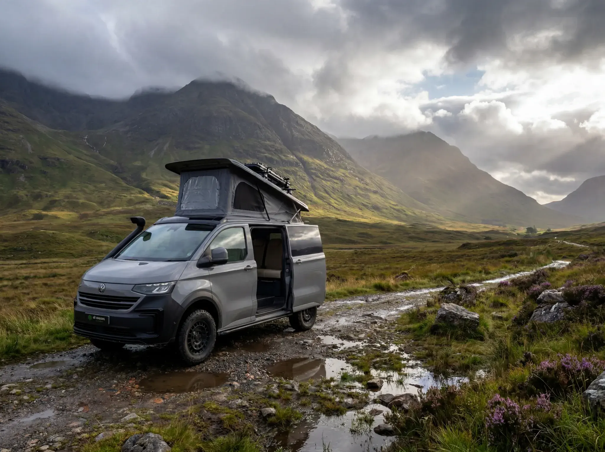 Silver VW Transporter campervan with pop-up roof parked on mountain track beside stream, Scottish Highlands backdrop.