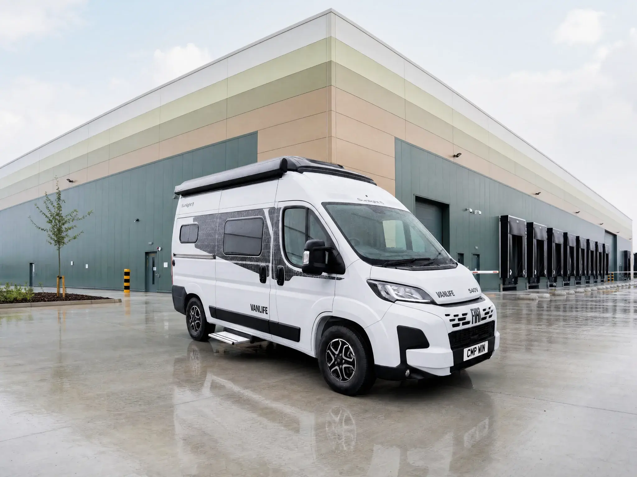 White Vanlife campervan parked on wet concrete forecourt outside modern warehouse building.