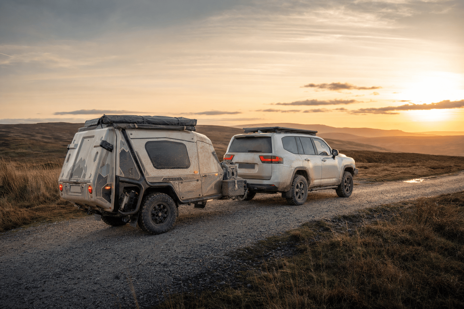 An off-road camper trailer towed behind a 4x4 parked on a gravel track across open British moorland at golden hour.