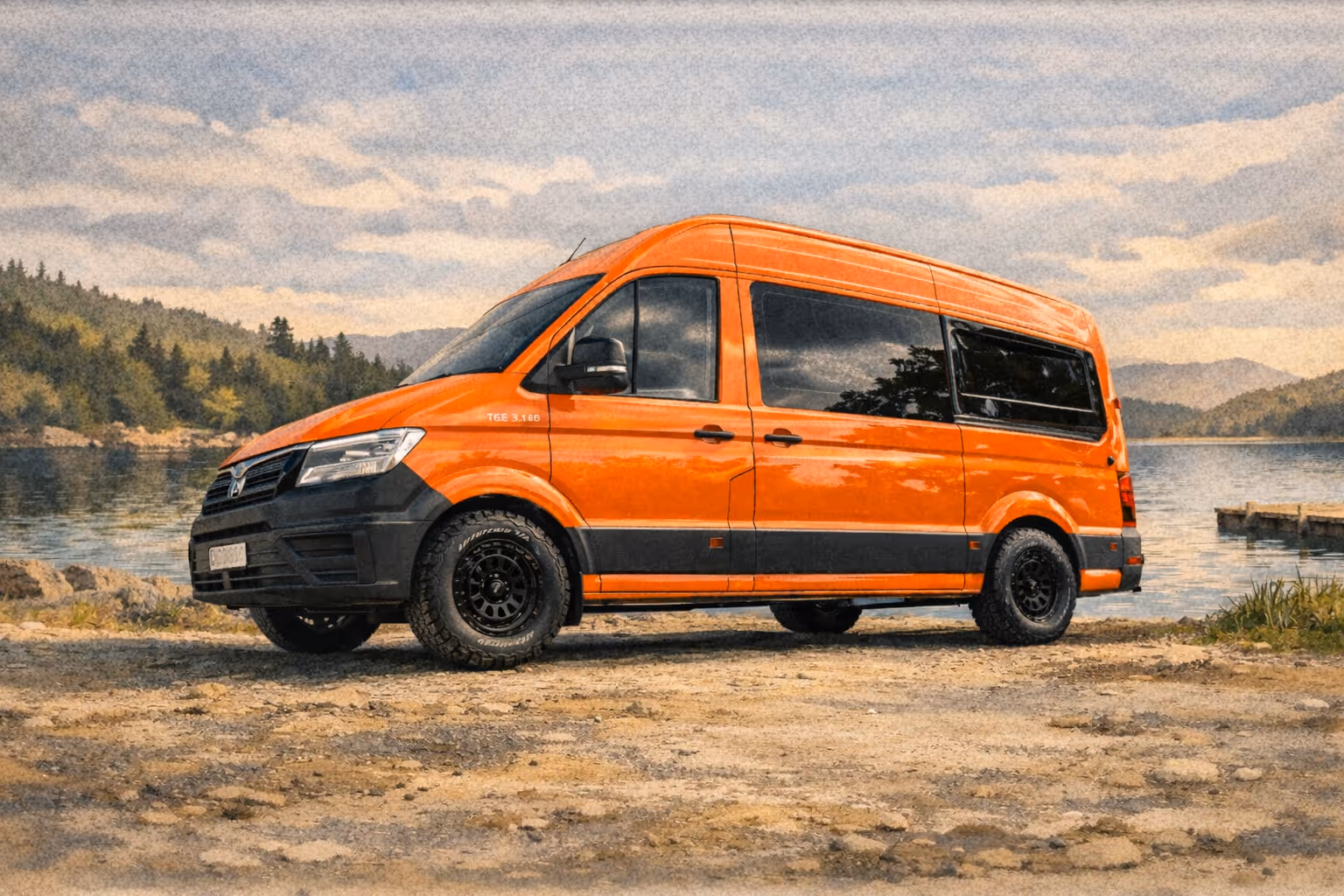 Orange and black campervan parked on gravel beside a lake with forested mountains in the background.
