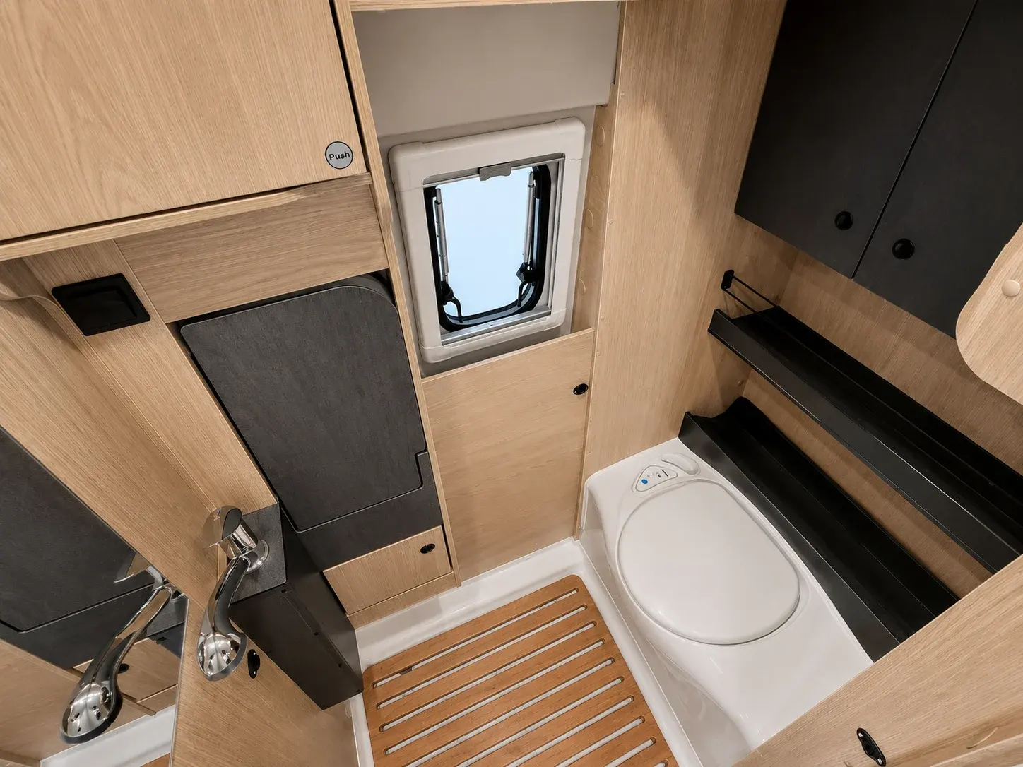 Campervan interior bathroom area with white toilet, wooden floor, window, and light wood cabinetry.