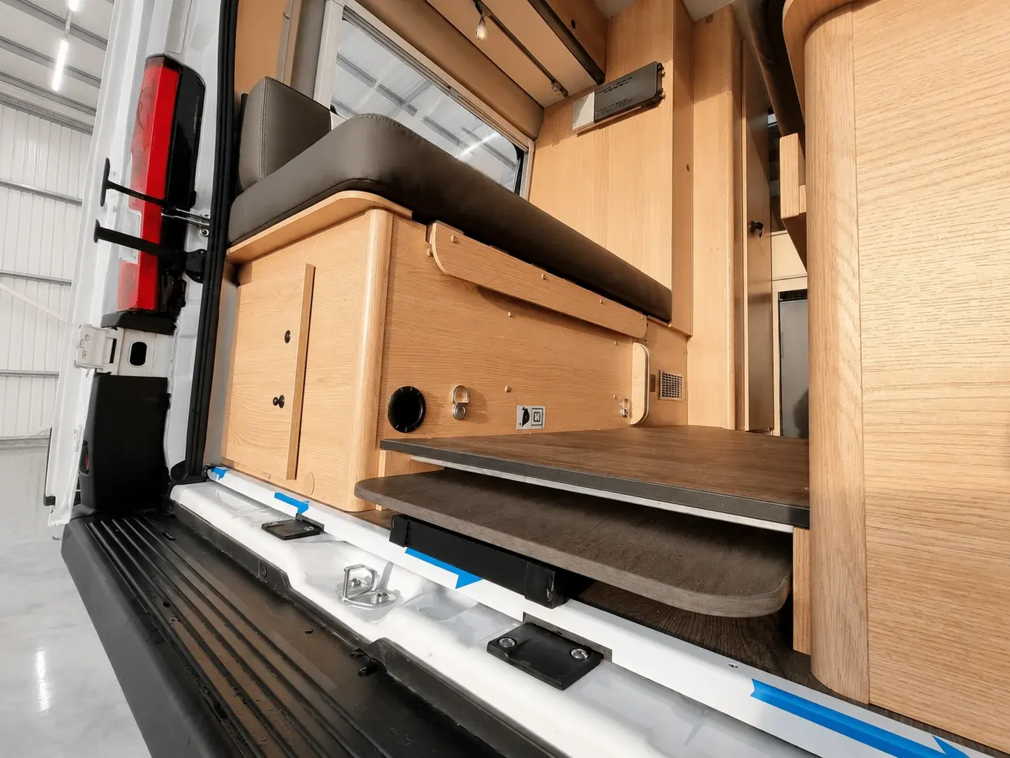 Campervan interior showing wooden cabinetry, pullout bed with dark cushion, and open rear doors with red tail lights.