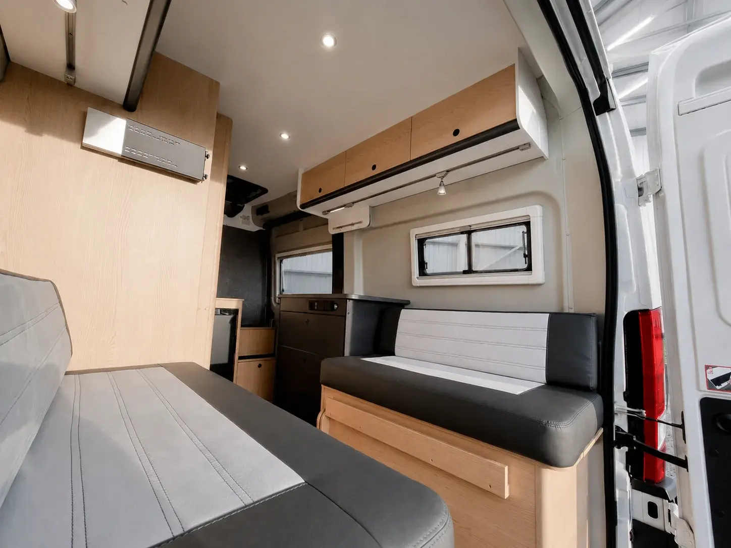 Campervan interior with wooden cabinetry, seating area with black and grey upholstery, and side windows.