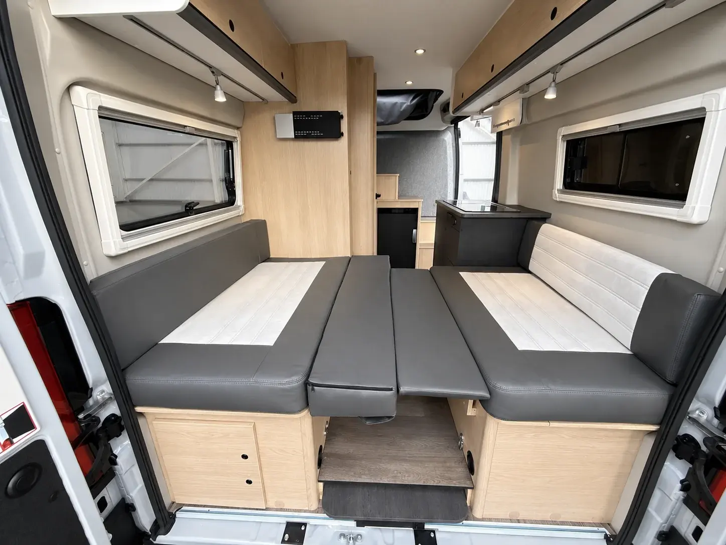 Campervan interior with grey upholstered bench seating, wooden cabinetry, windows, kitchenette, and convertible bed setup.