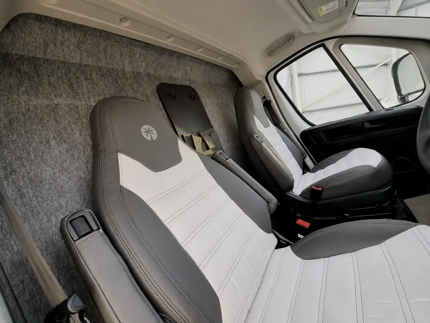 Campervan interior with grey and white upholstered front seats, fabric headlining, and windows showing storage area.