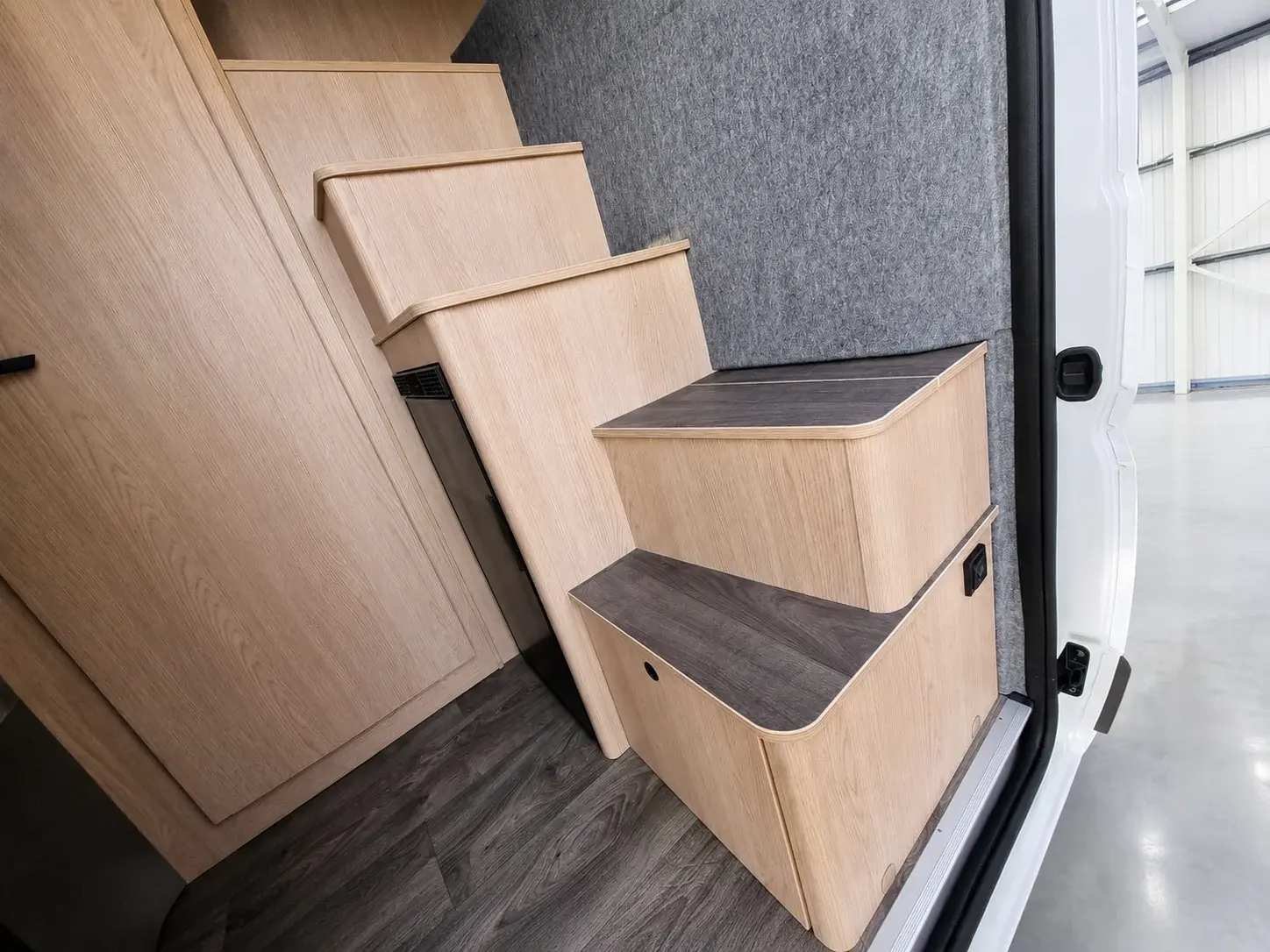 Campervan interior with light wood cabinetry, grey upholstered wall panel, and two-step wooden storage stairs with dark surfaces.