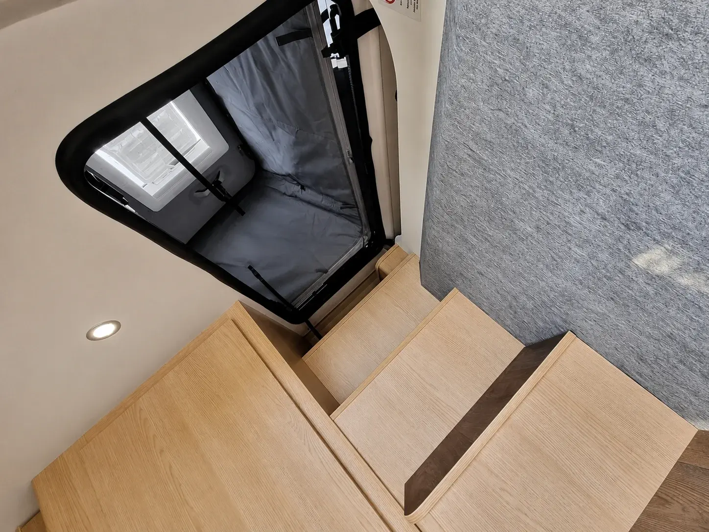 Campervan interior with wooden flooring, gray upholstered seating, and open black window blind with skylight visible.