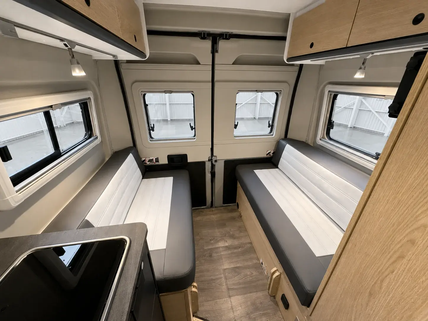 Campervan interior with dual bunk beds, wooden cabinetry, multiple windows, and light wood flooring.