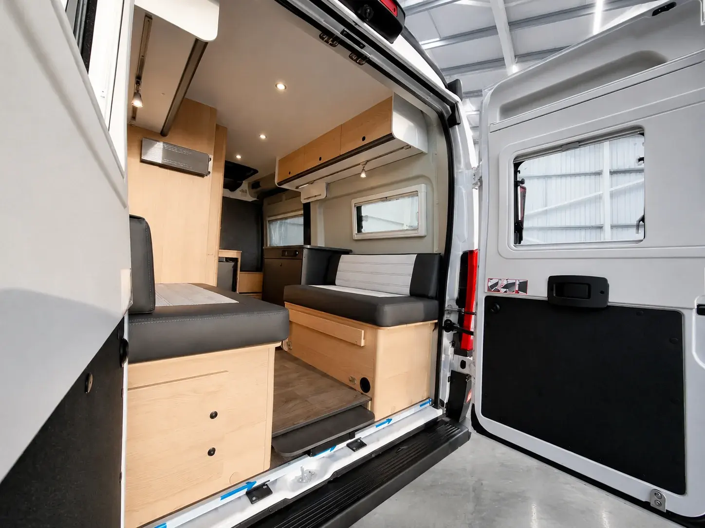 White campervan interior with wooden cabinetry, black upholstered seating, windows, and kitchen area visible through open rear doors.