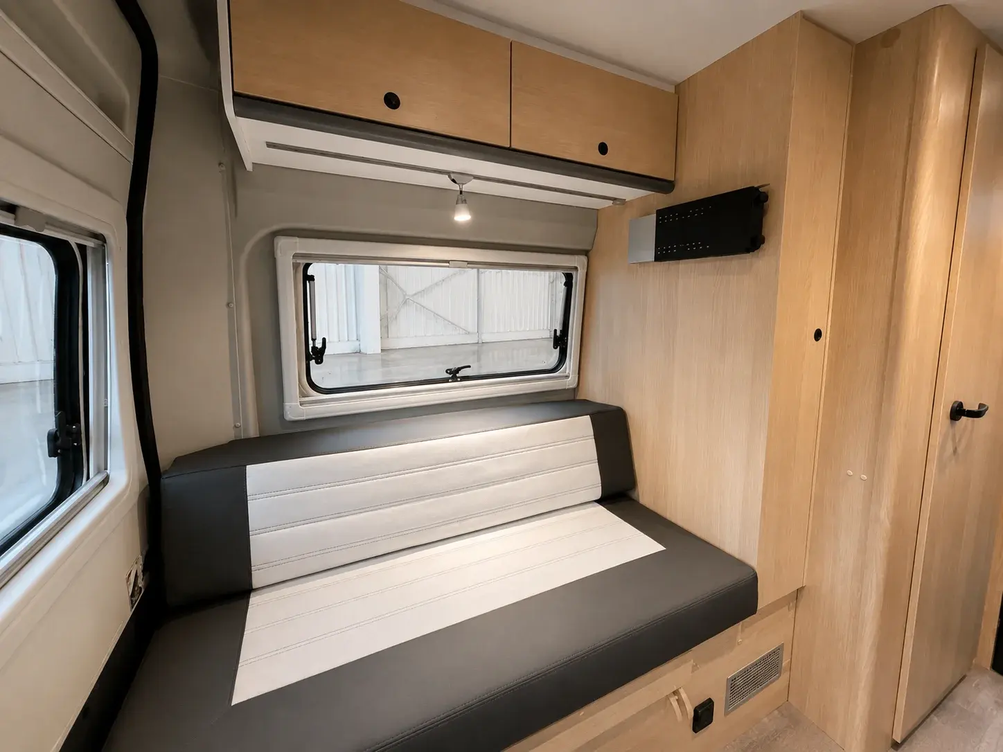 Campervan interior with wooden cabinetry, black and white seating bench, and window overlooking industrial building.