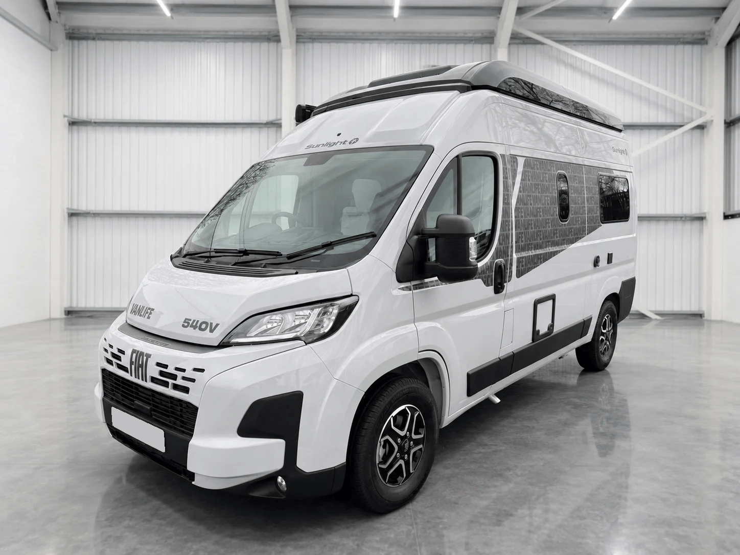 White Fiat-based campervan with black roof rack and solar panels, parked in bright warehouse.