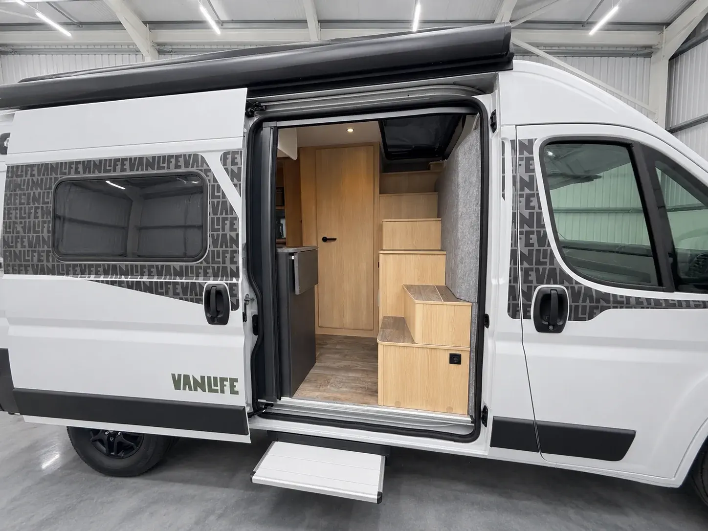 White campervan with open rear door revealing wooden interior steps and light wood cabinetry.